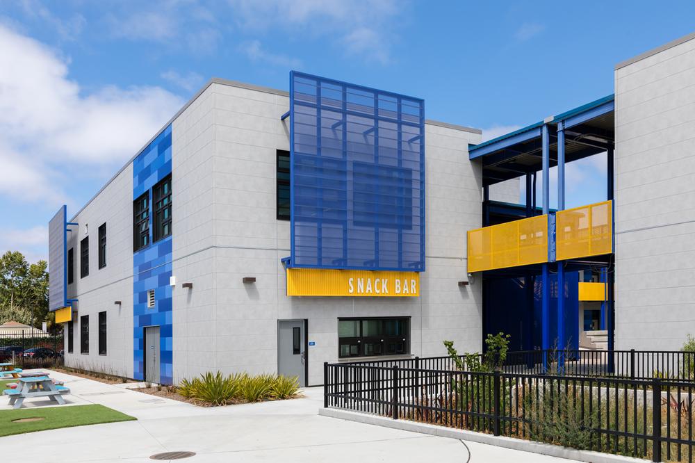The exterior of a school building with gray fiber cement siding accented with sections of blue and yellow signage and railings.