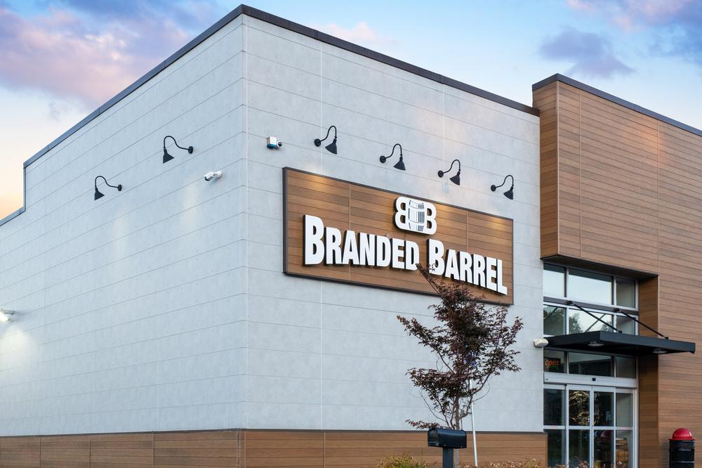 The front of a Branded Barrel store utilizing gray and cedar fiber cement siding. Black lamps illuminated the logo.