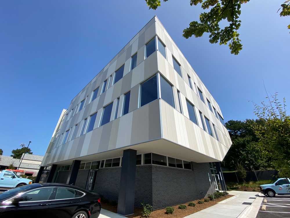 A commercial building has white, gray and tan siding. There are two blue trucks and a black car parked in the lot.