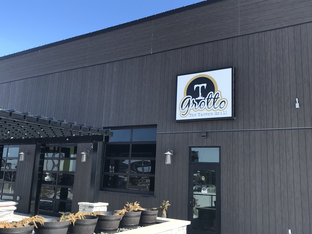 A restaurant with wood siding on the exterior, black windows, a pergola and planters out front. 