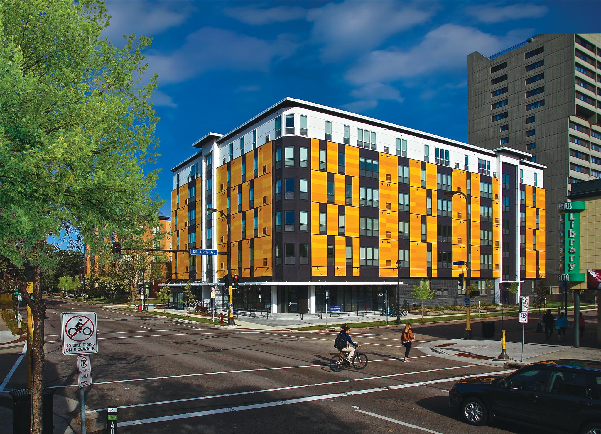 A biker and pedestrian passing the multi-story student housing with yellow exterior.