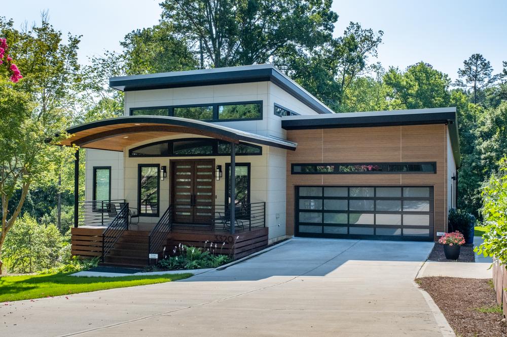This modern house has an attached garage with wood-like paneling. The front porch has black chairs on it.