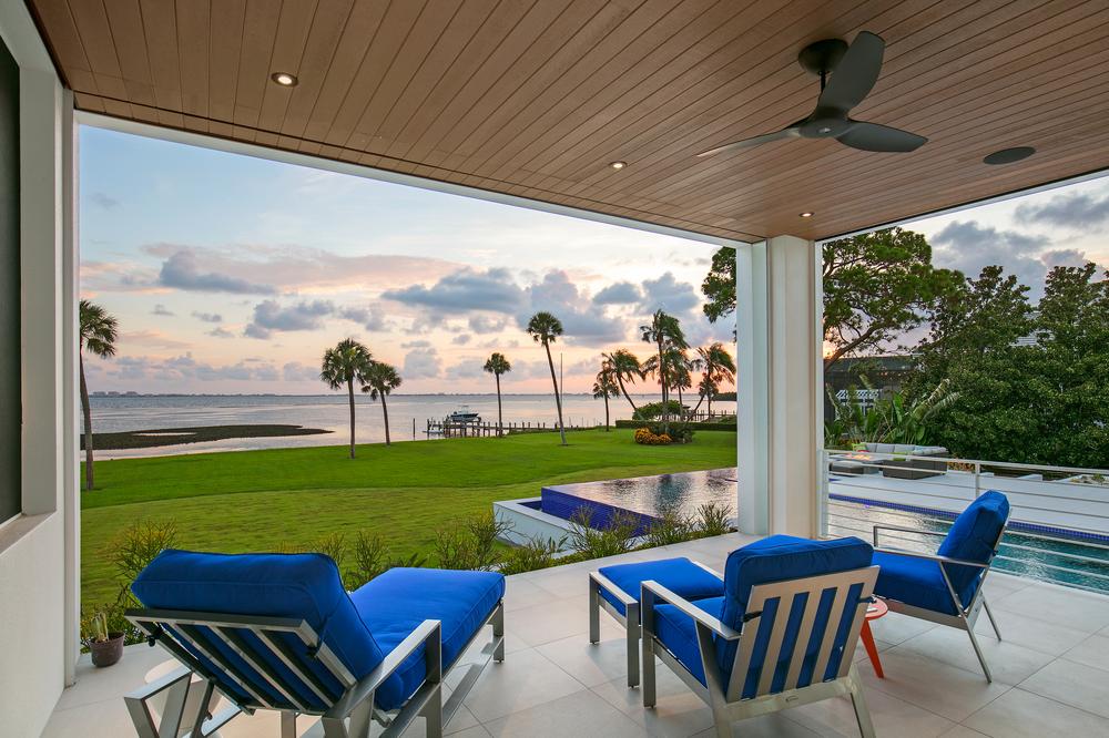 View of the ocean and palm trees from a backyard patio on the bottom floor of a coastal home. Blue chairs are arranged on the patio near a pool in the backyard.