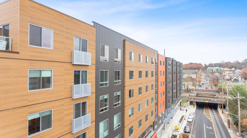 This student housing building features fiber cement siding. There is a road that is beside the building, and there are cars parked next to the sidewalk.
