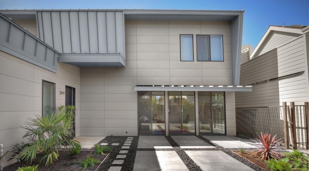 The backyard of a modern two-story home with sliding glass doors, beige siding and an asymmetrical gray roof.
