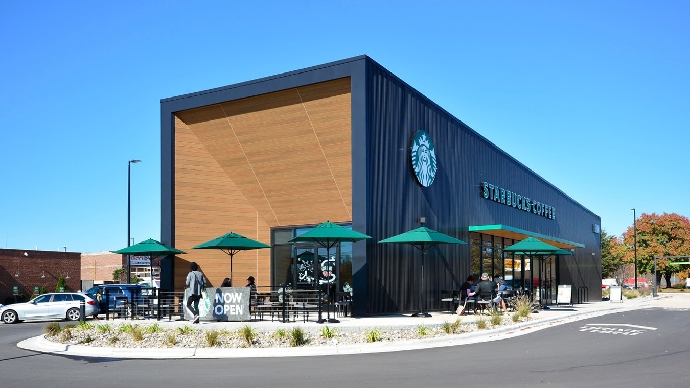 Exterior of Starbucks location featuring angled soffit and wall products. There are green umbrellas at the outdoor seating areas and a “Now Open” sign out front.