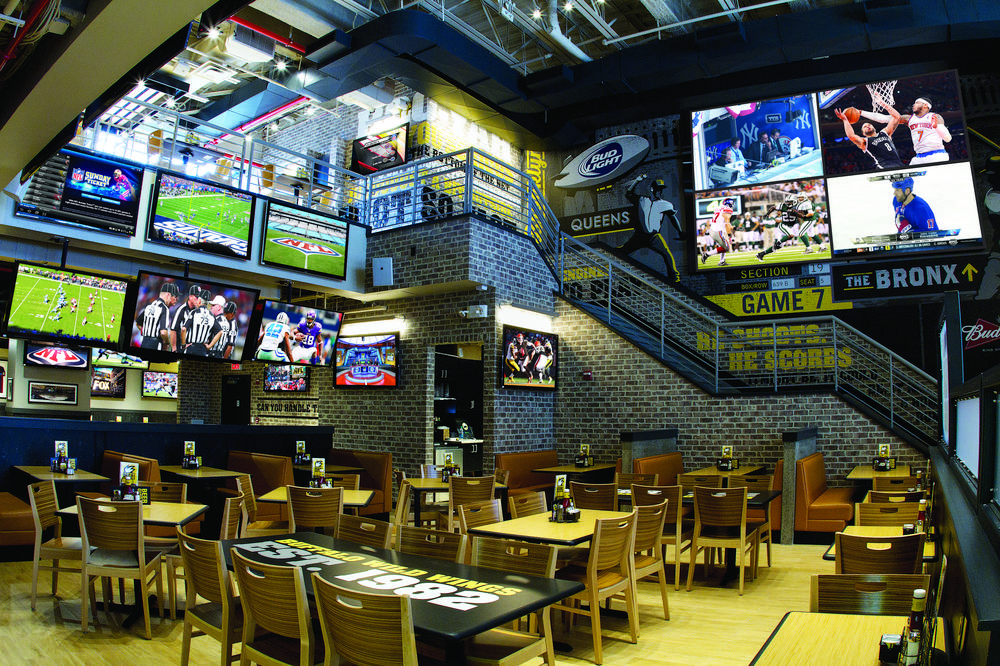 The interior of a two-story sports bar with light brick interior. The walls are covered with posters and television screens.