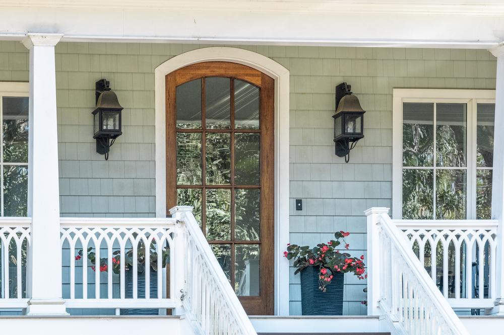 This close-up image shows a home’s front porch. There are fiber cement cedar shakes in a light blue color. There are two lights on the sides of the wooden front door, and there are two flower pots below the lights.