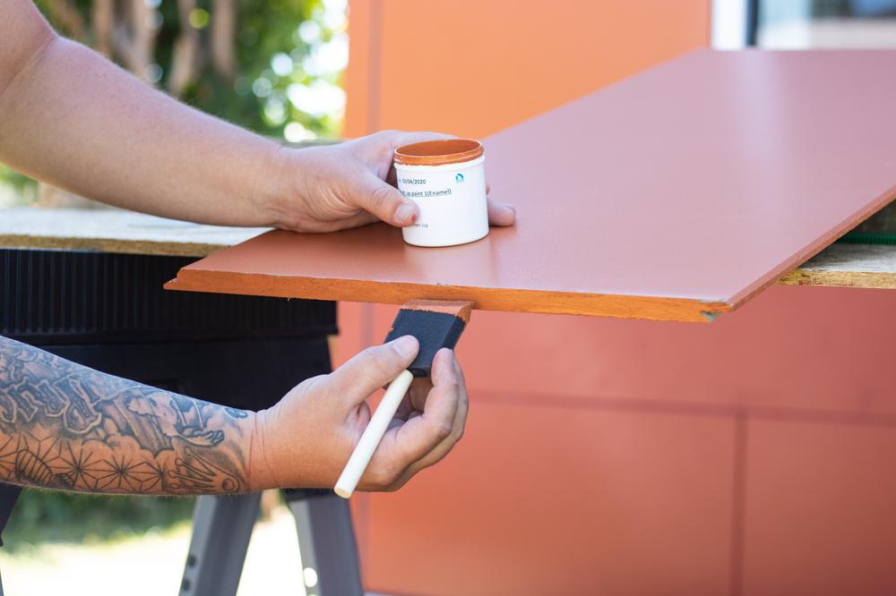 A hand using a foam paint brush to paint Nichiha Illumination fiber cement panels orange.