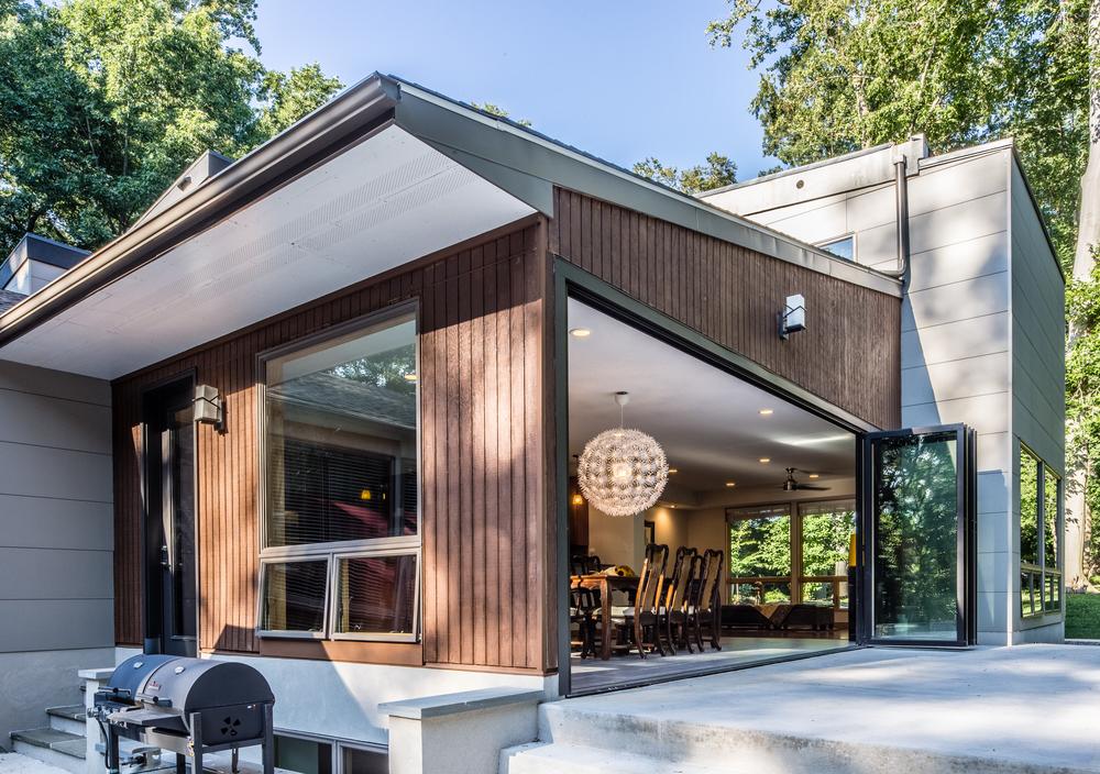 A home decorated with vertical, brown Nichiha fiber cement siding with the back door open, a table highlighted under a chandelier, and a grill outside.